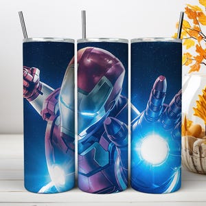 May include: A cylindrical metal tumbler featuring a graphic of a red and gold armored character in a space setting. The character has glowing blue eyes and a bright blue light emanating from the palm of a hand. A thin metal straw is inserted at the top.