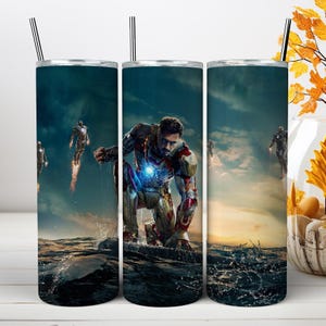 May include: A cylindrical stainless steel tumbler featuring a wrap-around graphic of Iron Man in a red and gold suit kneeling on water. The design includes a dark, cloudy sky background, glowing blue chest arc reactor, and flying figures.