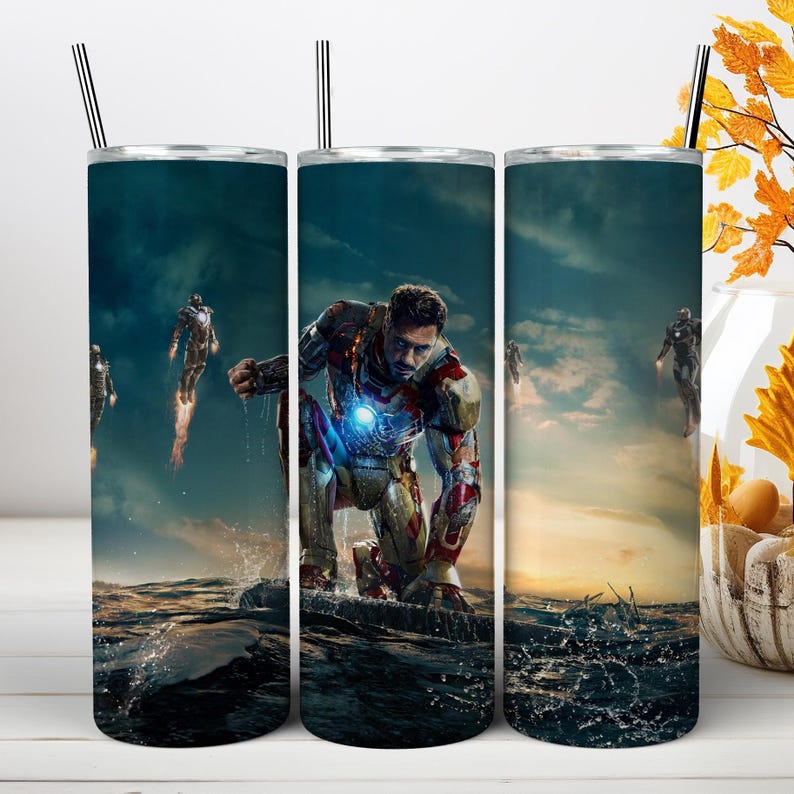 May include: A cylindrical stainless steel tumbler featuring a wrap-around graphic of Iron Man in a red and gold suit kneeling on water. The design includes a dark, cloudy sky background, glowing blue chest arc reactor, and flying figures.