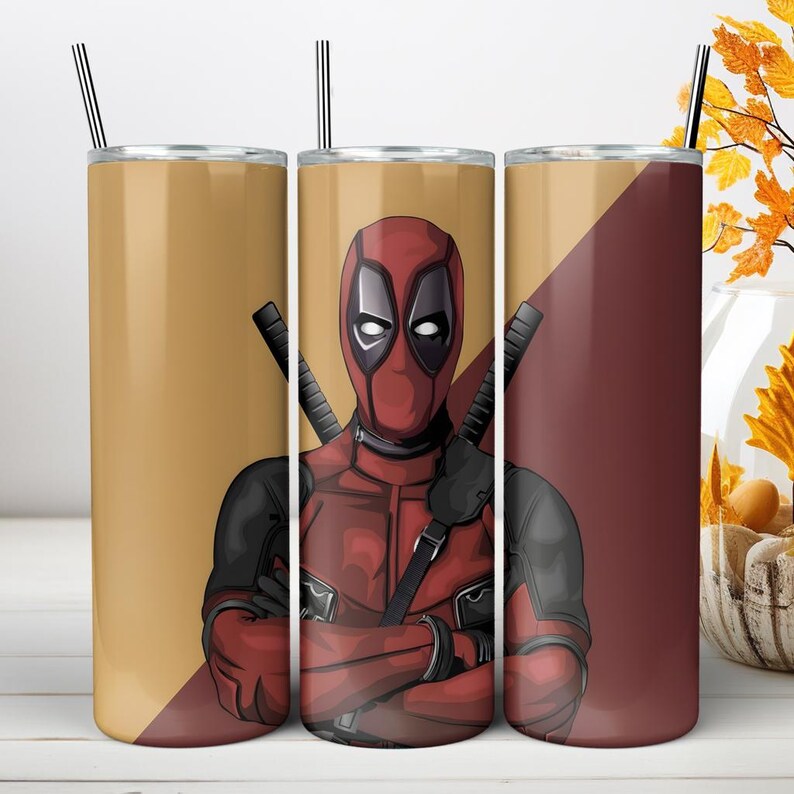 May include: A cylindrical tumbler featuring a graphic illustration of the character Deadpool in a red and black suit with crossed swords on the back. The design includes a split background of tan and dark red, topped with a thin metal straw.