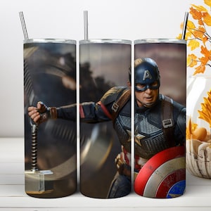 May include: A cylindrical metal tumbler featuring a graphic print of Captain America in a blue, red, and silver suit, holding a hammer and a round red, white, and blue shield. The tumbler includes a thin metal straw extending from the top lid.