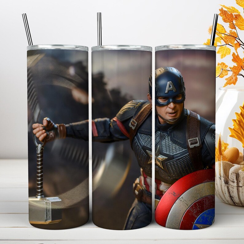 May include: A cylindrical metal tumbler featuring a graphic print of Captain America in a blue, red, and silver suit, holding a hammer and a round red, white, and blue shield. The tumbler includes a thin metal straw extending from the top lid.