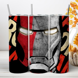 May include: A cylindrical tumbler with a straw featuring a graphic design of a red and silver armored mask with glowing white eyes. The surface has a distressed, textured appearance with bold red, black, and cream-colored geometric patterns.