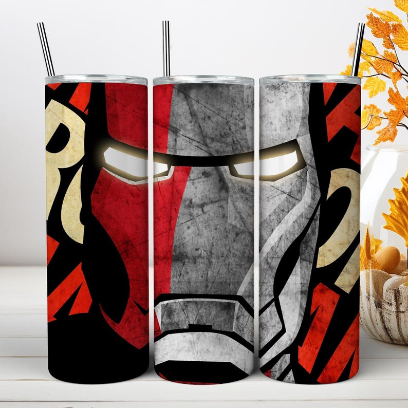 May include: A cylindrical tumbler with a straw featuring a graphic design of a red and silver armored mask with glowing white eyes. The surface has a distressed, textured appearance with bold red, black, and cream-colored geometric patterns.
