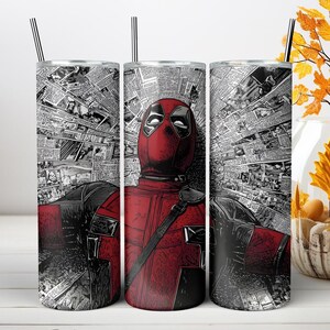 May include: A cylindrical stainless steel tumbler featuring a graphic of the character Deadpool in a red and black suit, set against a background of black and white newspaper clippings. The tumbler includes a thin metal straw inserted at the top.