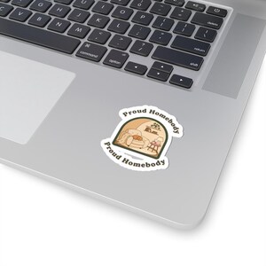 May include: A sticker with the text "Proud Homebody" in a curved design. The sticker features a cozy illustration of a living room scene with a sofa, a cat, and a small table. The sticker is placed on a laptop.