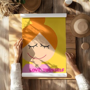 Könnte beinhalten: Gerollte Poster mit einer Cartoon-Illustration einer lächelnden Person mit geschlossenen Augen und orangefarbenem Haar vor gelbem Hintergrund. Der Text "LOVE YOURSELF" ist rosa. Das Poster liegt auf einer Holzoberfläche.