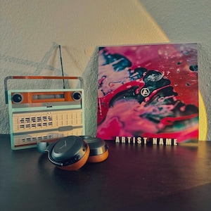 May include: A vintage-style radio with a light blue and brown finish, a pair of black and wood-toned headphones, and a colorful album cover with the text "ARTIST NAME" on a dark surface.
