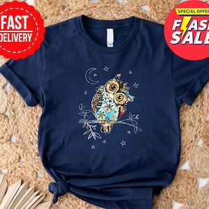 May include: Navy blue t-shirt featuring a colorful owl design perched on a branch, with a crescent moon and stars. The owl has large, detailed eyes and is adorned with floral and patterned elements. The shirt has a classic crew neck and short sleeves.