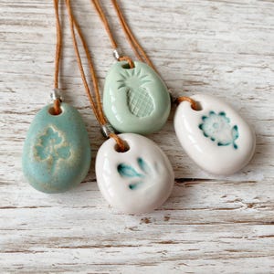 May include: Four ceramic pendant necklaces with brown cords. The oval pendants feature embossed designs: a four-leaf clover, a pineapple, a floral pattern, and a leaf design. The pendants are in shades of blue, green, and white.