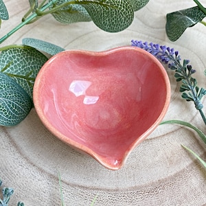 May include: A pink, heart-shaped ceramic dish with a small spout. The dish has a glossy finish and is set on a light-coloured wooden surface, with green leaves and purple flowers in the background. The dish is likely for serving small portions.