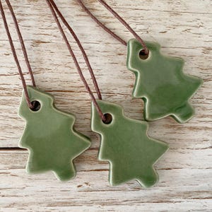 May include: Three green ceramic Christmas tree pendants on brown cords. The pendants have a glossy finish and are displayed against a light wood grain background. Each tree has a hole at the top for the cord.