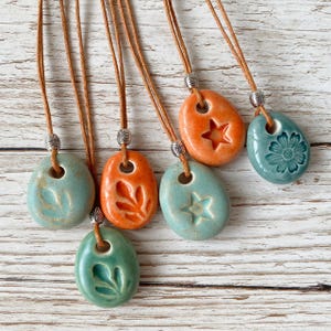 May include: Six ceramic pendant necklaces in shades of orange, teal, and green, each with a unique embossed design. The pendants are strung on brown cords with silver-colored metal accents. The designs include a star, leaf, and flower.