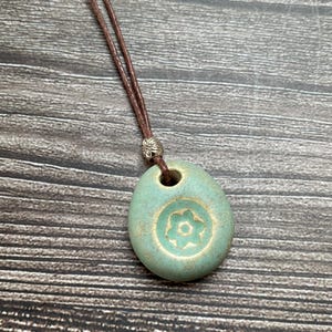 May include: A turquoise pendant necklace with a brown cord. The oval pendant features a flower design. The cord is attached with a silver bead. The necklace is a simple, elegant piece.