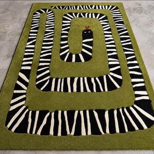 May include: A rectangular area rug featuring a solid olive green background with a large, coiled snake graphic. The snake has a black and white striped pattern across its body, a solid black head, and a small red tongue protruding from its mouth.