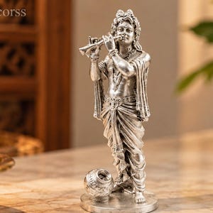 Könnte beinhalten: Eine silberfarbene Metallfigur einer stehenden Gottheit, die eine Flöte spielt. Die Figur trägt traditionelle drapierte Gewänder und Schmuck, mit einem kleinen kugelförmigen Gefäß zu den Füßen. Der Text Bohohomedecorss ist in der oberen Ecke sichtbar.