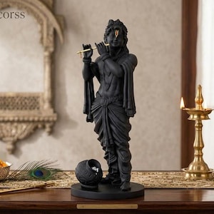 Puede incluir: Una estatua negra de Krishna tocando una flauta dorada, con un tilak dorado en la frente. La figura se alza sobre una base circular junto a una pequeña vasija negra. Dos lámparas de aceite de latón con llamas encendidas están a la derecha.