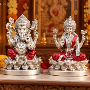 Könnte beinhalten: Zwei silberfarbene Statuen der Gottheiten Ganesha und Lakshmi, die auf Lotussockeln sitzen. Ganesha hat einen Elefantenkopf und vier Arme, während Lakshmi vier Arme hat und Lotusblumen hält. Beide Figuren tragen rote Gewänder und verzierte Kronen.