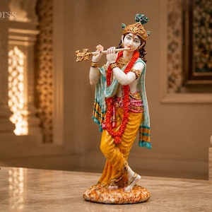 Könnte beinhalten: Eine stehende Krishna-Figur, die eine goldene Flöte spielt, mit weißem Teint, bekleidet mit einem gelben Dhoti, einer roten Blumengirlande und einer verzierten goldenen Krone mit Pfauenfeder-Akzent, auf einem strukturierten runden Sockel.