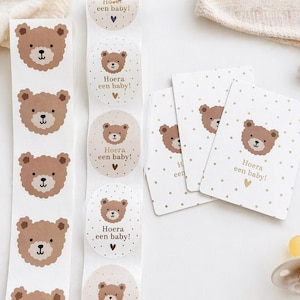 Op de afbeelding: Baby-thema artikelen, waaronder stickers en kaarten. De stickers en kaarten hebben een bruin teddybeerontwerp en de tekst "Hoera een baby!". Een fopspeen en een muts zijn ook zichtbaar.