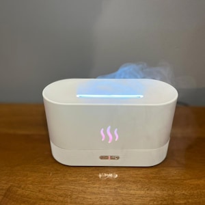 Puede incluir: Un difusor de aceites esenciales blanco, de forma ovalada, con una tira de luz azul y tres iconos de llama rosa. El difusor emite vapor. El dispositivo está sobre una superficie de madera.