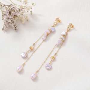 Puede incluir: Elegantes pendientes dorados con un delicado dise&ntilde;o de cadena. Los pendientes presentan racimos de perlas de color p&uacute;rpura claro y peque&ntilde;os detalles dorados. El dise&ntilde;o incluye una perla en forma de l&aacute;grima al final de la cadena.