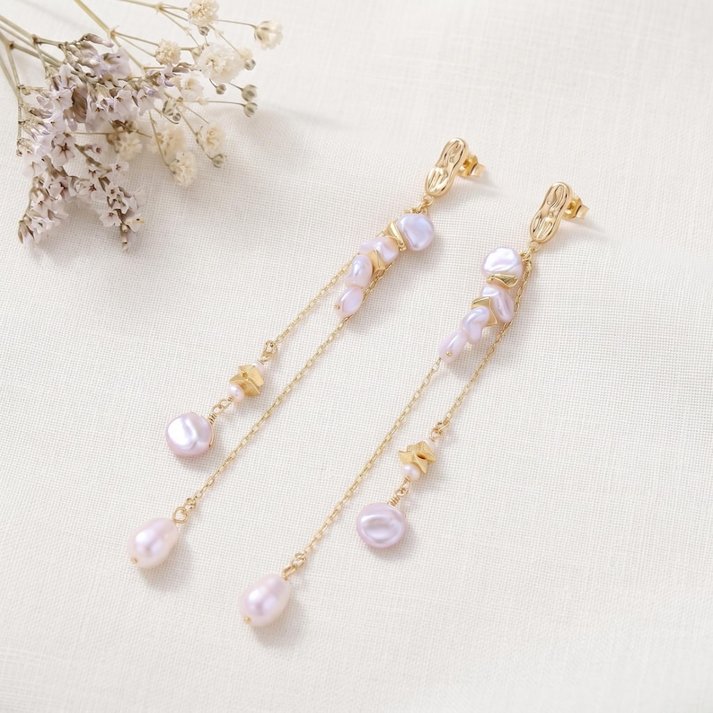 Puede incluir: Elegantes pendientes dorados con un delicado dise&ntilde;o de cadena. Los pendientes presentan racimos de perlas de color p&uacute;rpura claro y peque&ntilde;os detalles dorados. El dise&ntilde;o incluye una perla en forma de l&aacute;grima al final de la cadena.