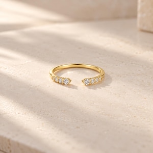 May include: A gold open-style ring featuring a polished band with a gap at the centre. Each end of the band is set with three small, round, clear, faceted stones that catch the light, creating a symmetrical design with a total of six stones.