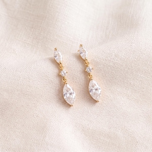 Pode incluir: Um par de brincos pendentes de ouro com uma pequena pedra redonda transparente no topo e uma pedra transparente maior de corte marquise na parte inferior, ligadas por uma fina corrente de ouro. Acabamento em ouro polido e pedras facetadas.