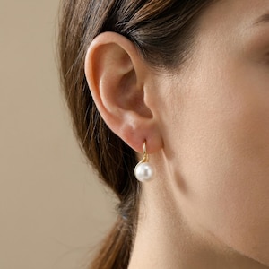 May include: A close-up of a gold earring with a white pearl. The earring is a simple hook design, with the pearl suspended below. The pearl is round and has a smooth, lustrous surface. The earring is worn on an ear.