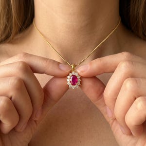 May include: A gold necklace with a pendant featuring a large, oval ruby surrounded by a halo of small, clear gemstones. The ruby is a vibrant red, and the necklace is held by a delicate gold chain.
