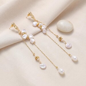 Puede incluir: Pendientes colgantes dorados con perlas blancas irregulares y perlas en forma de l&aacute;grima. Los pendientes tienen un delicado dise&ntilde;o de cadena con peque&ntilde;os detalles dorados. Los pendientes se muestran sobre una superficie color crema.
