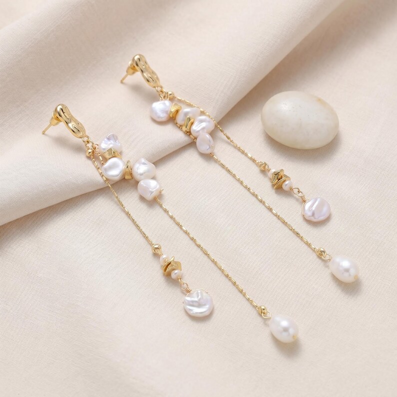 Puede incluir: Pendientes colgantes dorados con perlas blancas irregulares y perlas en forma de l&aacute;grima. Los pendientes tienen un delicado dise&ntilde;o de cadena con peque&ntilde;os detalles dorados. Los pendientes se muestran sobre una superficie color crema.