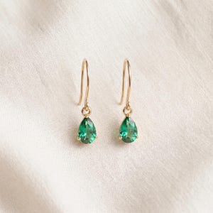 Può includere: Un paio di orecchini pendenti in oro con pietre preziose verdi sfaccettate a forma di goccia, sospese a semplici ganci dorati ricurvi. Gli orecchini presentano una finitura metallica lucida e una tonalità verde brillante e trasparente.