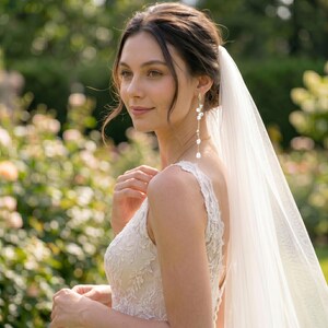 Puede incluir: Una mujer con un vestido de novia de encaje blanco y un velo transparente. Lleva pendientes largos dorados con detalles de perlas blancas. El fondo es un jard&iacute;n borroso con follaje verde y luz suave.