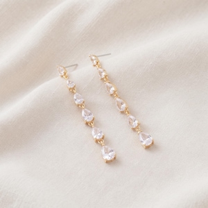 Peut inclure: Une paire de boucles d'oreilles pendantes dorées avec un design en chaîne verticale. Chaque boucle est ornée de quatre cristaux transparents à facettes en forme de poire disposés en ligne, suspendus à un fermoir à tige simple.