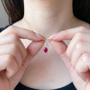 Puede incluir: Un collar de oro rosa con un colgante con una vibrante gema de rubí ovalada. El rubí está acentuado con pequeñas y brillantes gemas blancas. El collar está sostenido por una persona.
