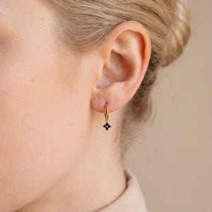 May include: A gold hoop earring featuring a small, black, four-petal flower pendant. The earring is displayed on a person's ear. The jewellery has a simple, elegant design.