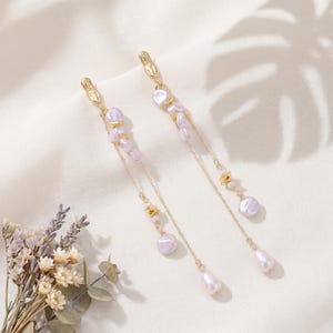Puede incluir: Pendientes dorados colgantes. Cada pendiente presenta perlas de color lila claro y peque&ntilde;os detalles dorados. Las cadenas terminan con una perla en forma de l&aacute;grima, creando un estilo elegante y sofisticado.