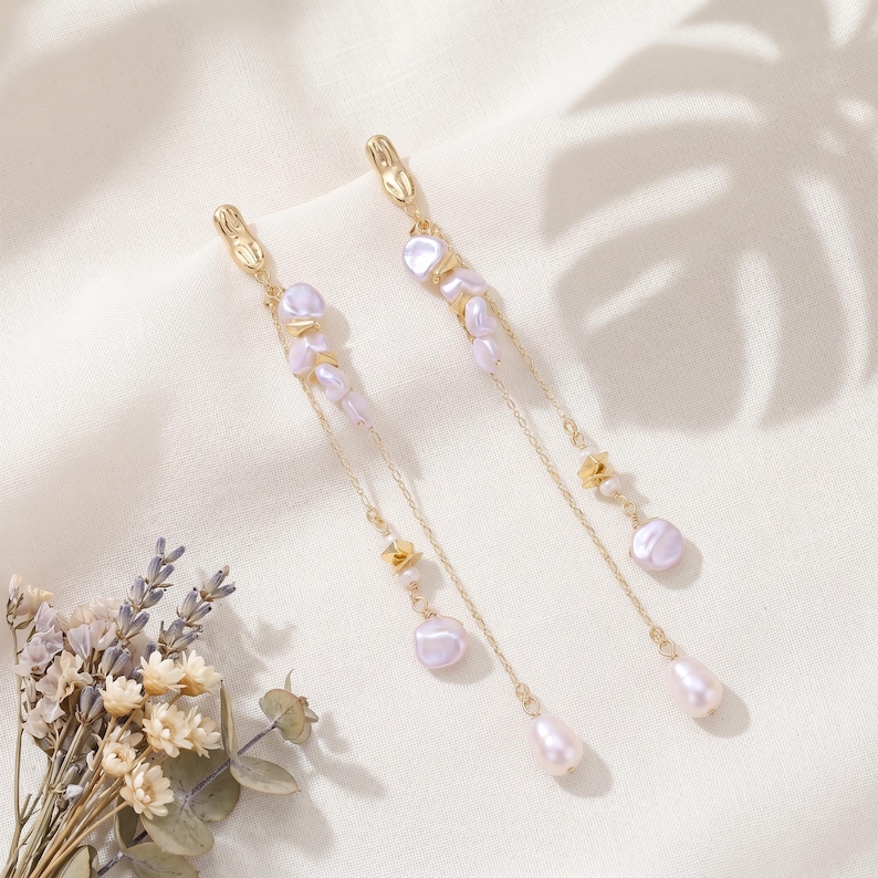 Puede incluir: Pendientes dorados colgantes. Cada pendiente presenta perlas de color lila claro y peque&ntilde;os detalles dorados. Las cadenas terminan con una perla en forma de l&aacute;grima, creando un estilo elegante y sofisticado.