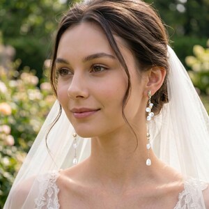Puede incluir: Una mujer lleva pendientes largos y colgantes con perlas blancas y peque&ntilde;as piedras transparentes. Los pendientes est&aacute;n sujetos a un metal dorado. Lleva un velo blanco y un vestido de encaje blanco. El fondo es un jard&iacute;n borroso.