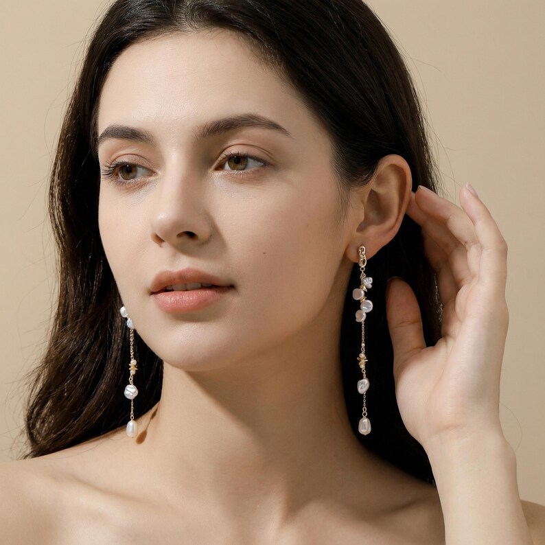 Puede incluir: Pendientes dorados con perlas blancas colgantes y peque&ntilde;os elementos decorativos. Los pendientes son largos y delgados, dise&ntilde;ados para colgar de los l&oacute;bulos de las orejas. Las perlas son de diferentes tama&ntilde;os, lo que a&ntilde;ade inter&eacute;s visual al dise&ntilde;o.