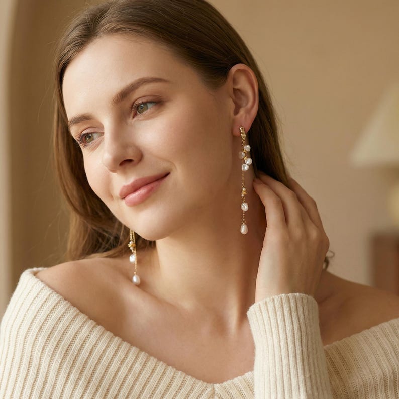 Puede incluir: Elegantes pendientes dorados con perlas blancas colgantes. Los pendientes presentan un dise&ntilde;o delicado con m&uacute;ltiples perlas que caen en cascada desde el l&oacute;bulo de la oreja. La mujer de la imagen lleva un jersey color crema.