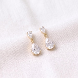 May include: A pair of gold-tone drop earrings featuring a small round clear crystal stud connected to a larger pear-shaped clear crystal pendant. The metal settings are thin and polished, holding the faceted stones securely in place.