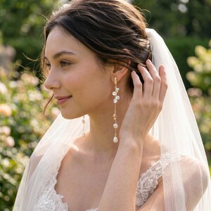 Puede incluir: Pendientes largos dorados con perlas colgantes y delicados detalles florales. La persona lleva un vestido de encaje blanco y un velo, lo que sugiere un tema nupcial. Los pendientes son de dise&ntilde;o delicado, adecuados para ocasiones formales.