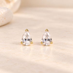 May include: A pair of stud earrings featuring pear-shaped clear stones set in gold-toned metal. Each stone is secured by three small prongs at the base and a single prong at the pointed top, creating a teardrop silhouette with a polished finish.