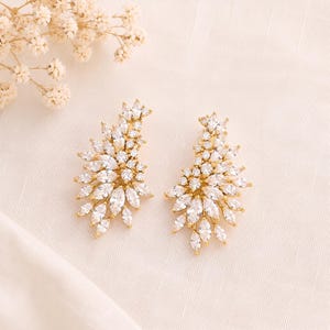 May include: A pair of gold-toned earrings featuring a cluster of clear, marquise-cut and round-cut crystals arranged in a fan-like, asymmetrical shape. The crystals are set in a metallic frame, creating a bright, reflective surface on each earring.