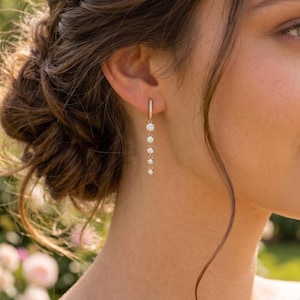 May include: A gold-toned drop earring featuring a small hinged hoop at the top, connected to a vertical chain of five round, clear, faceted stones that decrease slightly in size as they descend, creating a delicate and linear dangling design.