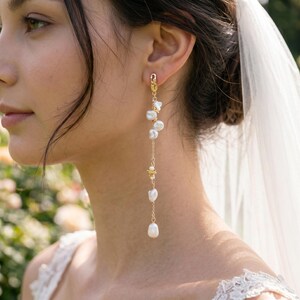Puede incluir: Un pendiente largo dorado con una serie de perlas blancas de forma irregular y peque&ntilde;os detalles florales. El pendiente cuelga de una fina cadena. Lo lleva una persona con cabello oscuro y un velo de novia blanco.