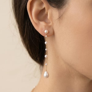 May include: Dangling pearl earrings with a gold chain. The earrings feature a small pearl stud at the ear, with a series of pearls descending in size, culminating in a larger, teardrop-shaped pearl at the bottom. The earrings are worn on a person.